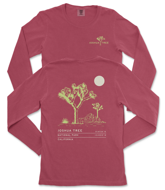 a red long-sleeved t-shirt with a graphic design depicting a desert landscape, including a tree and a moon. The text on the t-shirt reads "JOSUA TREE" and "33.8747° N 115.9013° W", indicating the location of Joshua Tree National Park in California.